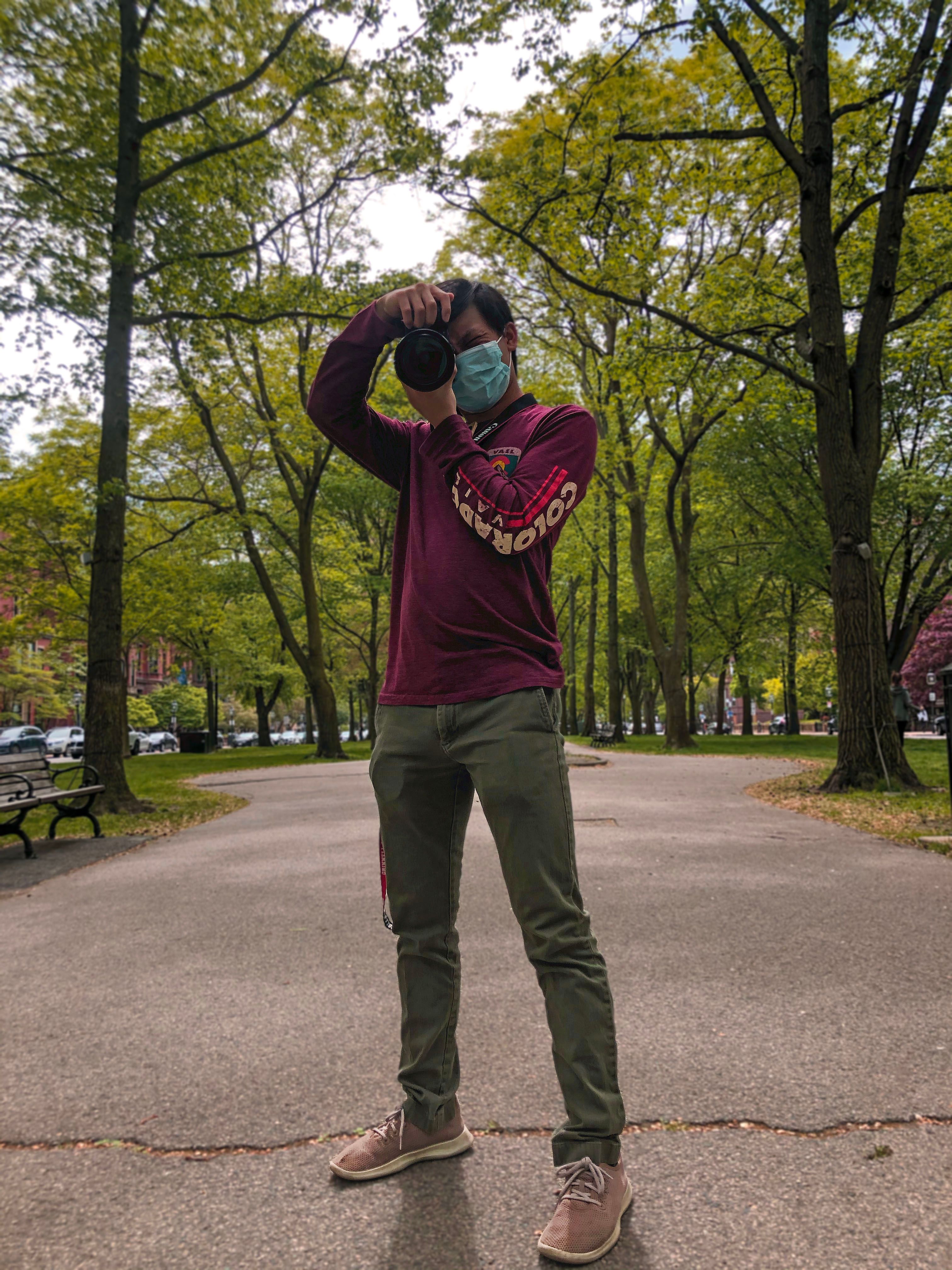 Masked photographer aiming a DSLR at the viewer on a tree-lined park promenade